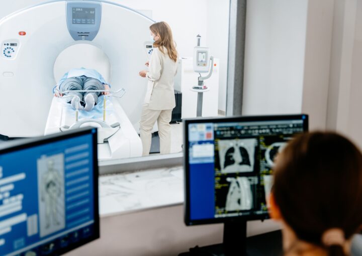 Patient liegt in CT-Röhre, medizinisches Personal daneben; im Vordergrund Monitore mit CT-Bildern.