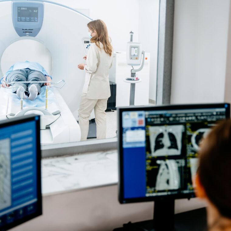Patient liegt in CT-Röhre, medizinisches Personal daneben; im Vordergrund Monitore mit CT-Bildern.
