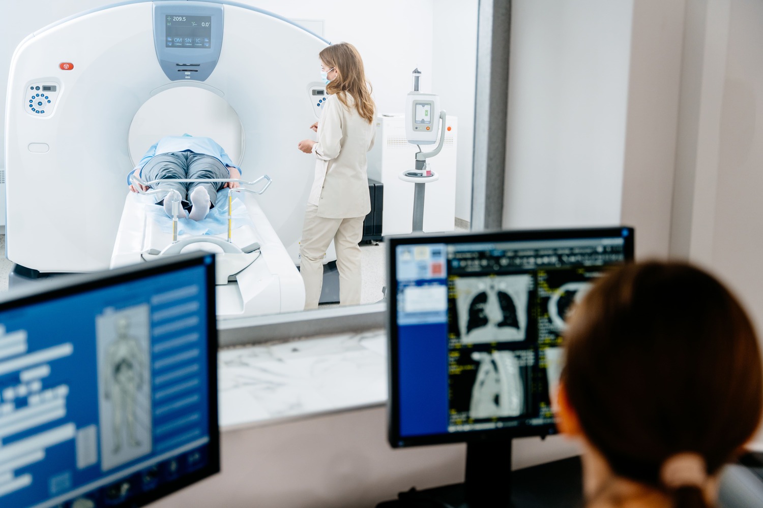 Patient liegt in CT-Röhre, medizinisches Personal daneben; im Vordergrund Monitore mit CT-Bildern.