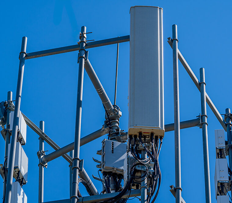 Nahaufnahme eines Mobilfunkmasts mit mehreren Antennen und Technikmodulen vor blauem Himmel. Zahlreiche Kabel verlaufen zu den Antenneneinheiten.