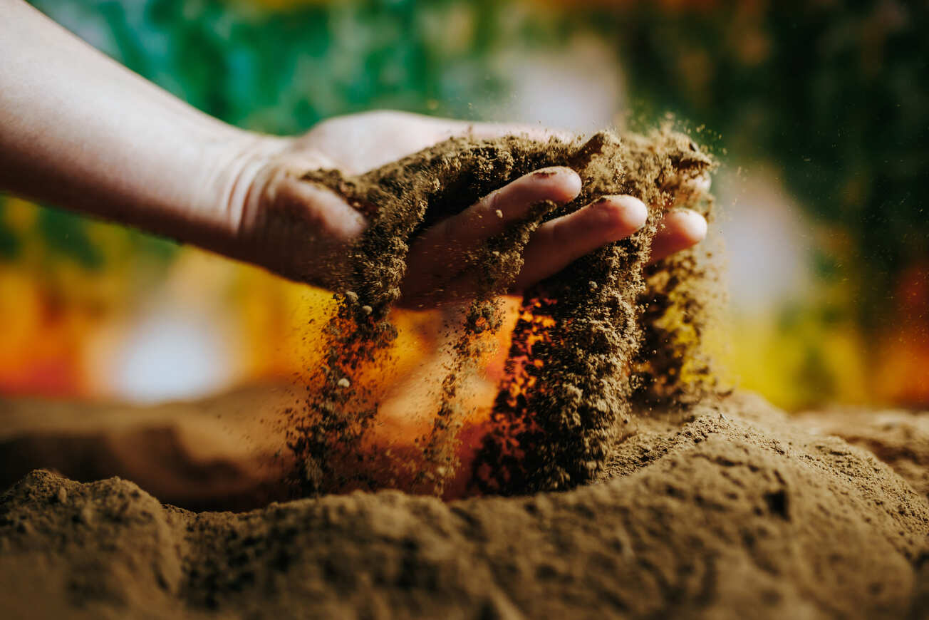 Hand lässt trockene Erde durch die Finger rieseln, bunter Hintergrund.