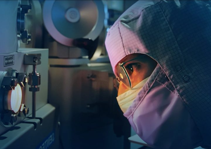 Person in Reinraumkleidung und Schutzbrille blickt konzentriert durch ein Sichtfenster an einem Hightech-Gerät in einem Labor.