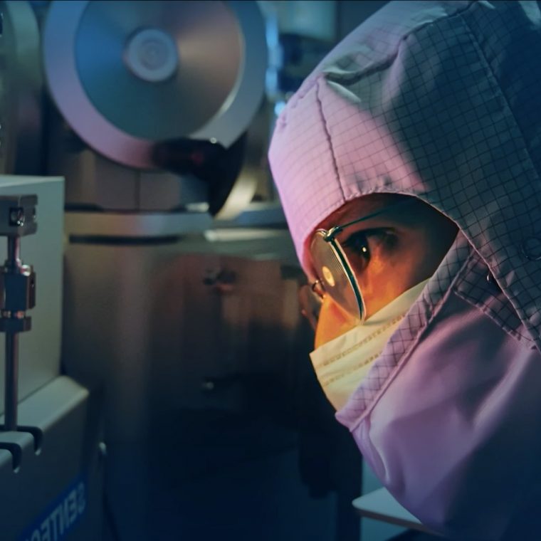 Person in Reinraumkleidung und Schutzbrille blickt konzentriert durch ein Sichtfenster an einem Hightech-Gerät in einem Labor.