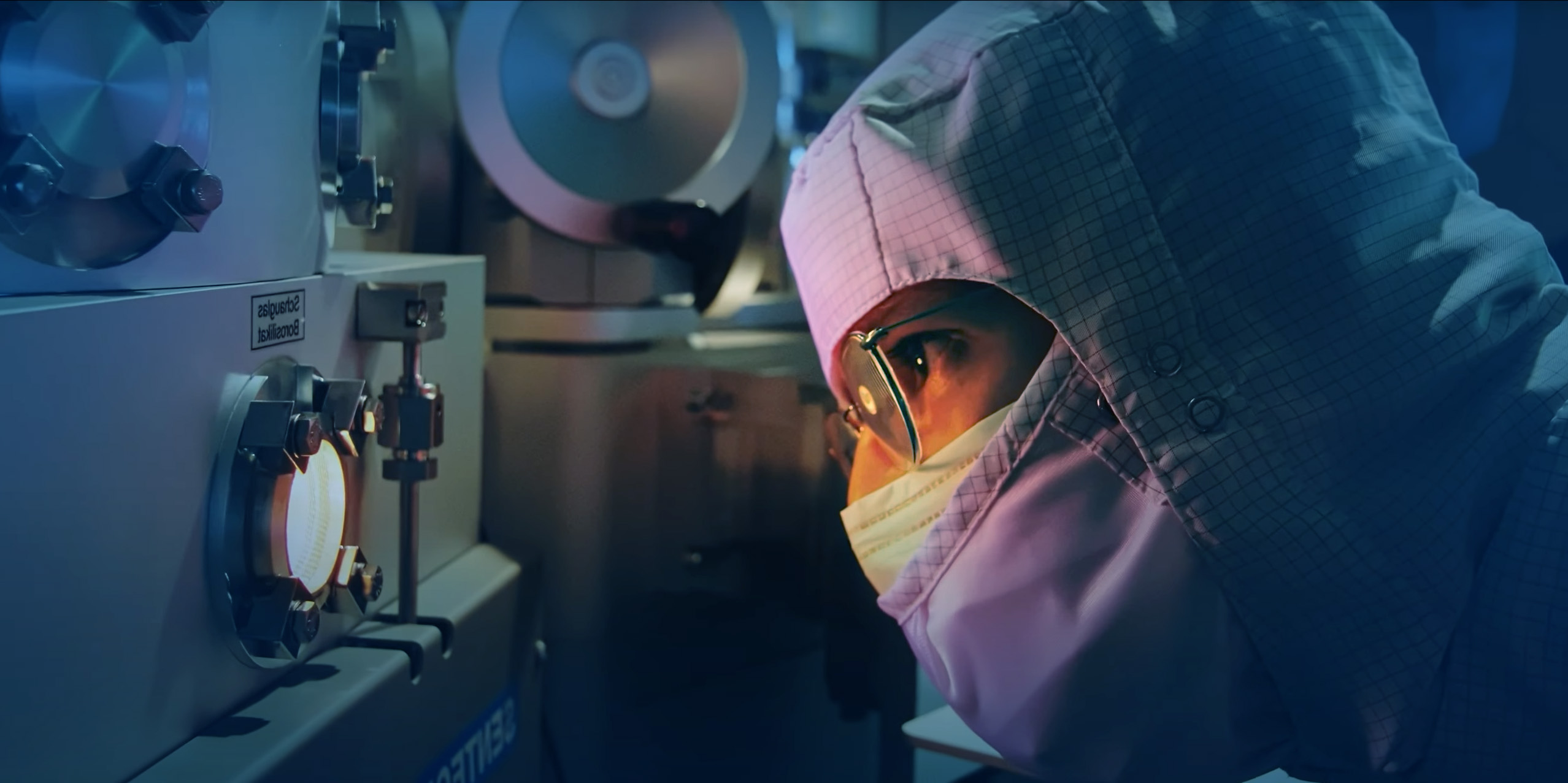 Person in Reinraumkleidung und Schutzbrille blickt konzentriert durch ein Sichtfenster an einem Hightech-Gerät in einem Labor.
