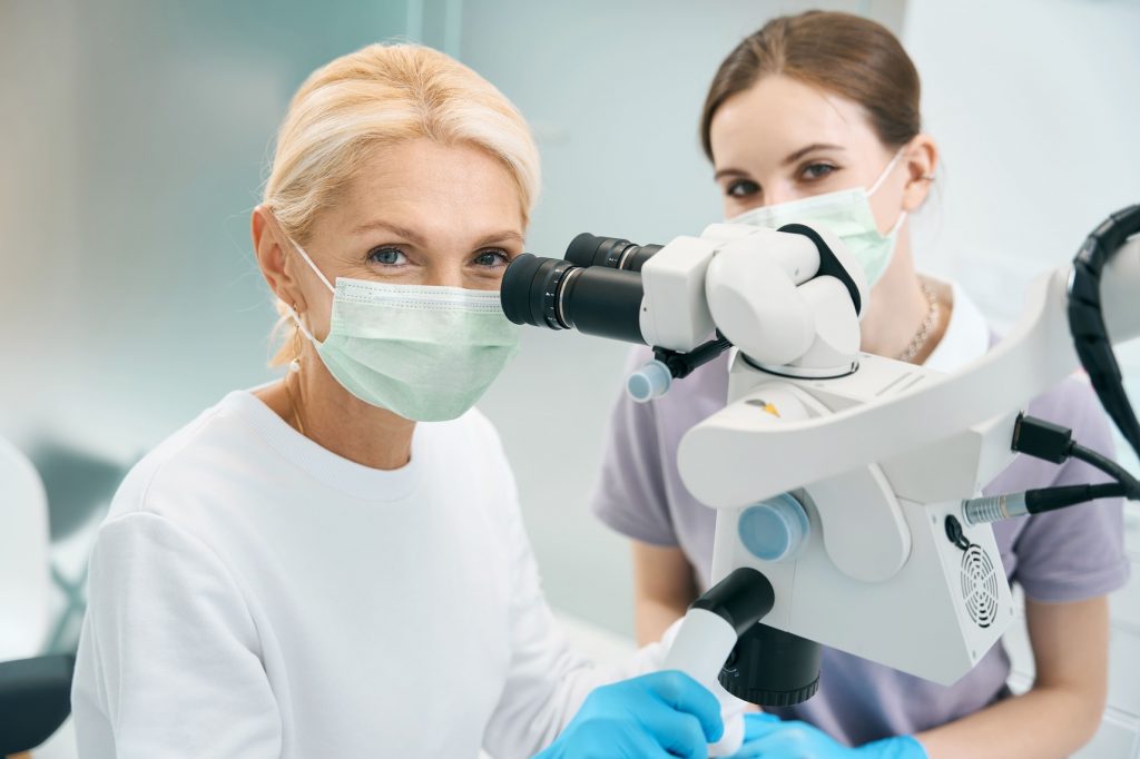 Zwei Forscherinnen mit Mundschutz betrachten eine Probe durch ein modernes Labormikroskop; Beide Frauen schauen direkt in die Kamera.