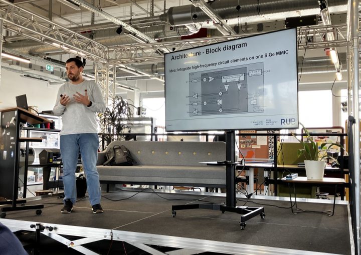 Mann hält einen technischen Vortrag auf einer kleinen Bühne in einem modernen Raum mit offener Decke; auf dem Bildschirm hinter ihm ist ein Blockdiagramm mit dem Titel ‚Architecture – Block diagram‘ zu sehen, darunter Logos von RUB und Viventes Systeme.