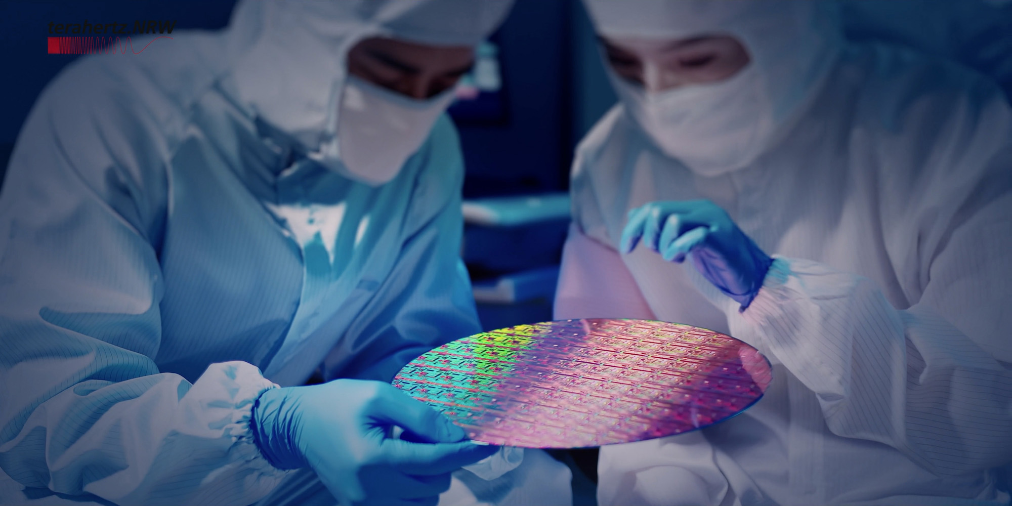Zwei Personen in Reinraumkleidung und blauen Handschuhen untersuchen einen glänzenden, bunten Silizium-Wafer; oben links ist das Logo von terahertz.NRW sichtbar.