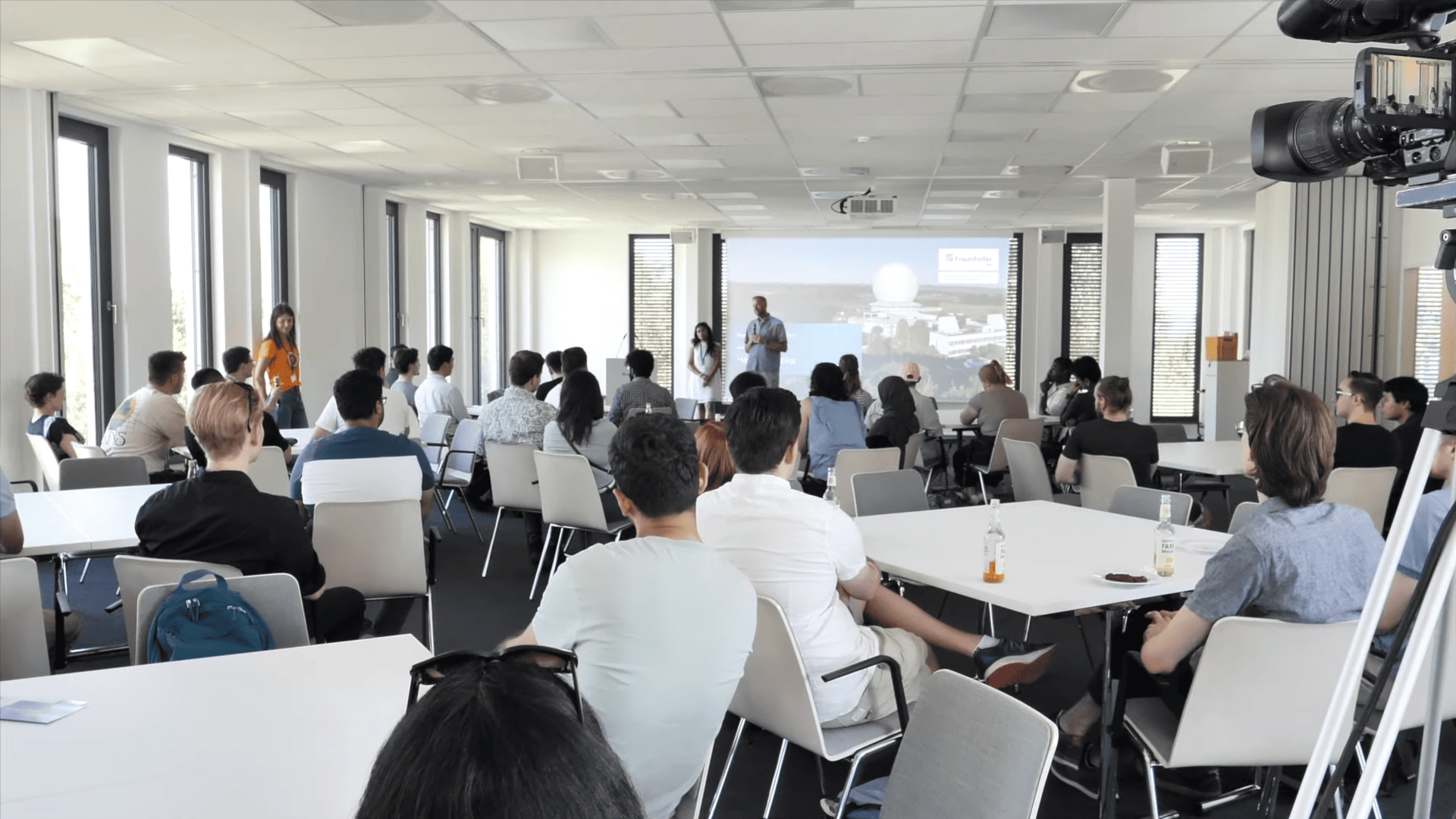 Menschen sitzen in einem modernen Konferenzraum und hören einem Vortrag zu, der vorne mit Präsentationsfolie gehalten wird; eine Kamera filmt die Veranstaltung.