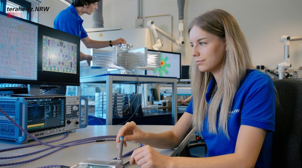 Junge Frau in blauem terahertz.NRW-Shirt arbeitet konzentriert an einem Laborarbeitsplatz mit technischen Geräten und Monitoren; im Hintergrund bedient ein Kollege ein Messgerät.