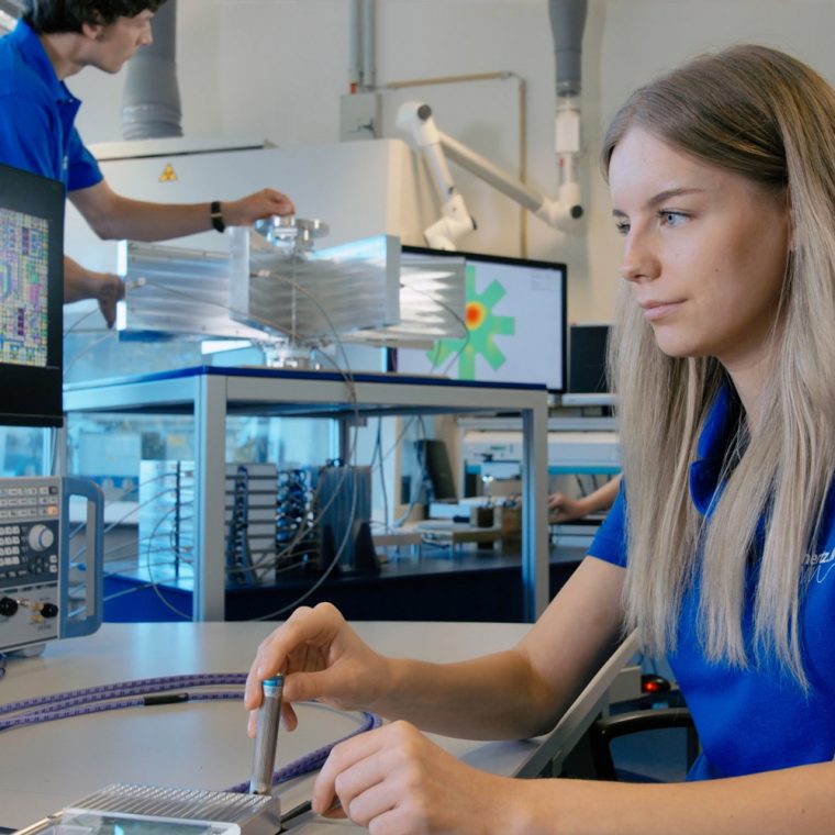 Junge Frau in blauem terahertz.NRW-Shirt arbeitet konzentriert an einem Laborarbeitsplatz mit technischen Geräten und Monitoren; im Hintergrund bedient ein Kollege ein Messgerät.