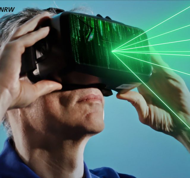 Mann mit grauen Haaren trägt eine Virtual-Reality-Brille, aus der grüne Laserstrahlen austreten; im Hintergrund das Logo von terahertz.NRW.