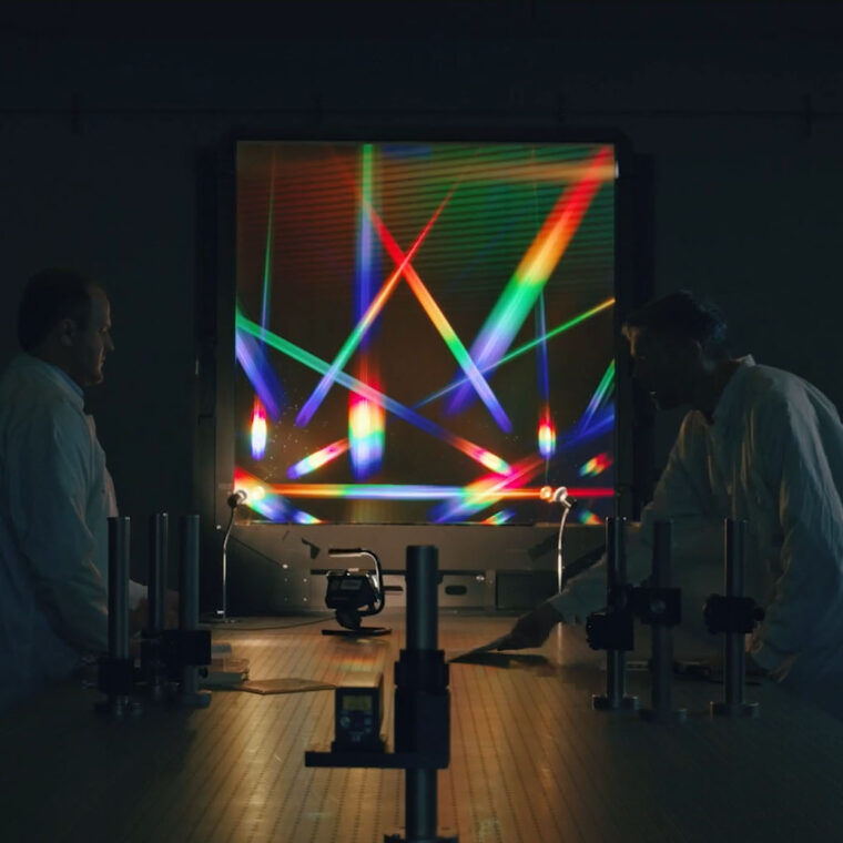 Zwei Forschende im Labor vor einer leuchtenden optischen Experimentieranlage mit farbigen Lichtstrahlen.