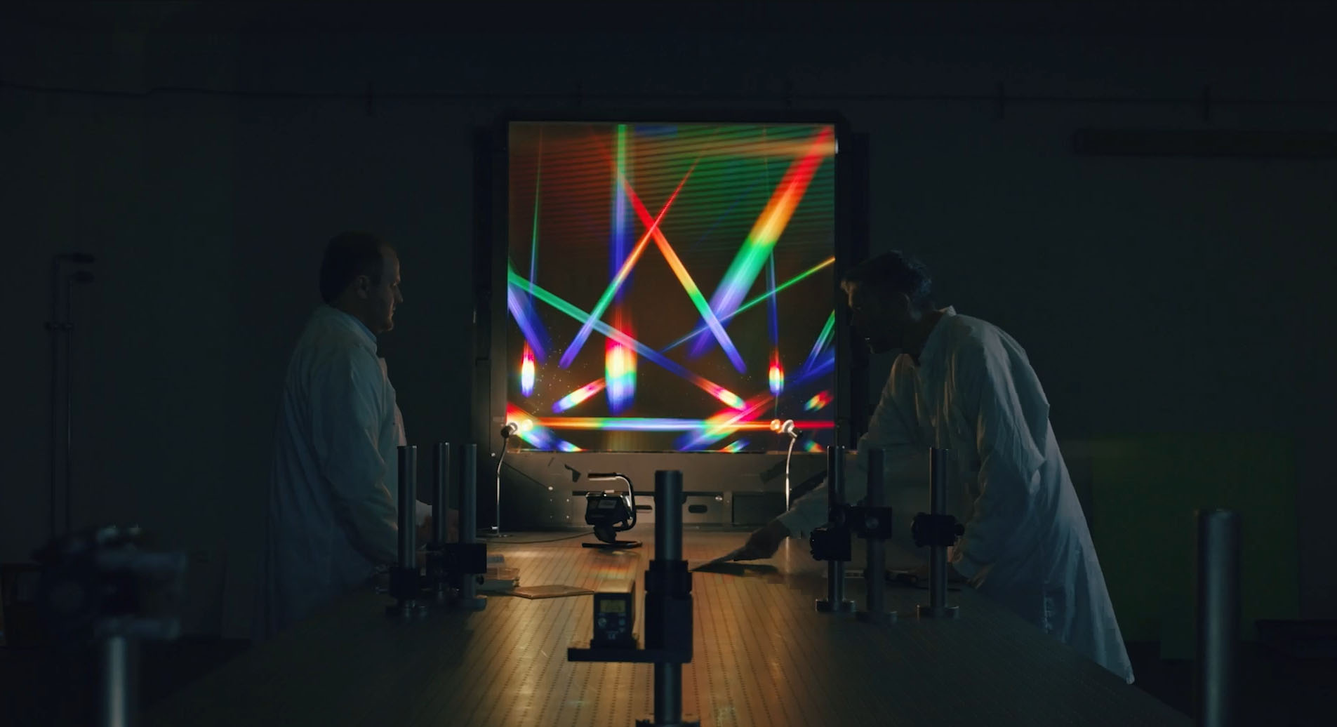 Zwei Forschende im Labor vor einer leuchtenden optischen Experimentieranlage mit farbigen Lichtstrahlen.