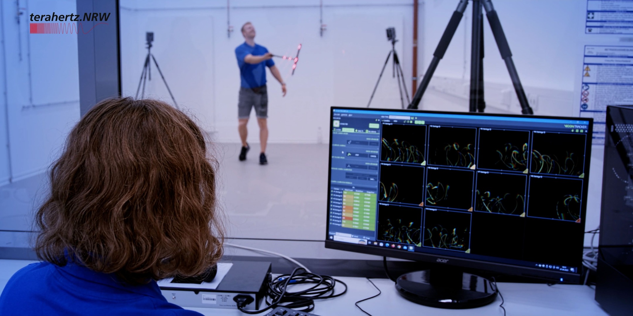 Eine Frau beobachtet durch eine Glasscheibe eine Bewegungserfassung. Im Hintergrund jongliert ein Mann mit einem LED-markierten Objekt, während Kameras die Bewegung aufzeichnen. Auf dem Monitor vor der Frau sind mehrere grafische Bewegungsanalysen sichtbar. Oben links ist das Logo von terahertz.NRW.