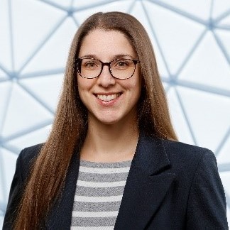 Hanne Bendel im Portrait vor einer geometrisch strukturierte Glas- oder Stahlkonstruktion. Sie trägt ein dunkelblaues Sakko über einem gestreiften Oberteil.