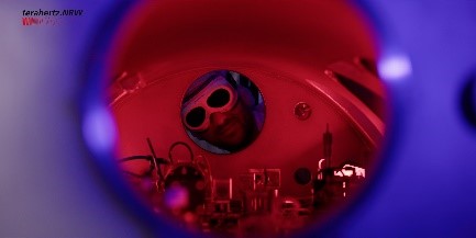 Blick ins Innere eines wissenschaftlichen Geräts mit rotem Licht. Eine Person mit Schutzbrille schaut durch ein rundes Sichtfenster. Im Inneren sind elektronische und optische Komponenten zu erkennen. Oben links ist das Logo von terahertz.NRW sichtbar.
