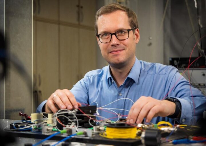 Ein Mann mit Brille und blauem Hemd sitzt an einem Labortisch mit elektronischen Komponenten und vielen Kabeln. Er blickt lächelnd in die Kamera und arbeitet konzentriert an einem Versuchsaufbau.