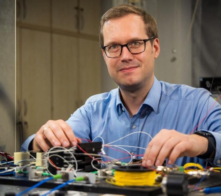 Ein Mann mit Brille und blauem Hemd sitzt an einem Labortisch mit elektronischen Komponenten und vielen Kabeln. Er blickt lächelnd in die Kamera und arbeitet konzentriert an einem Versuchsaufbau.