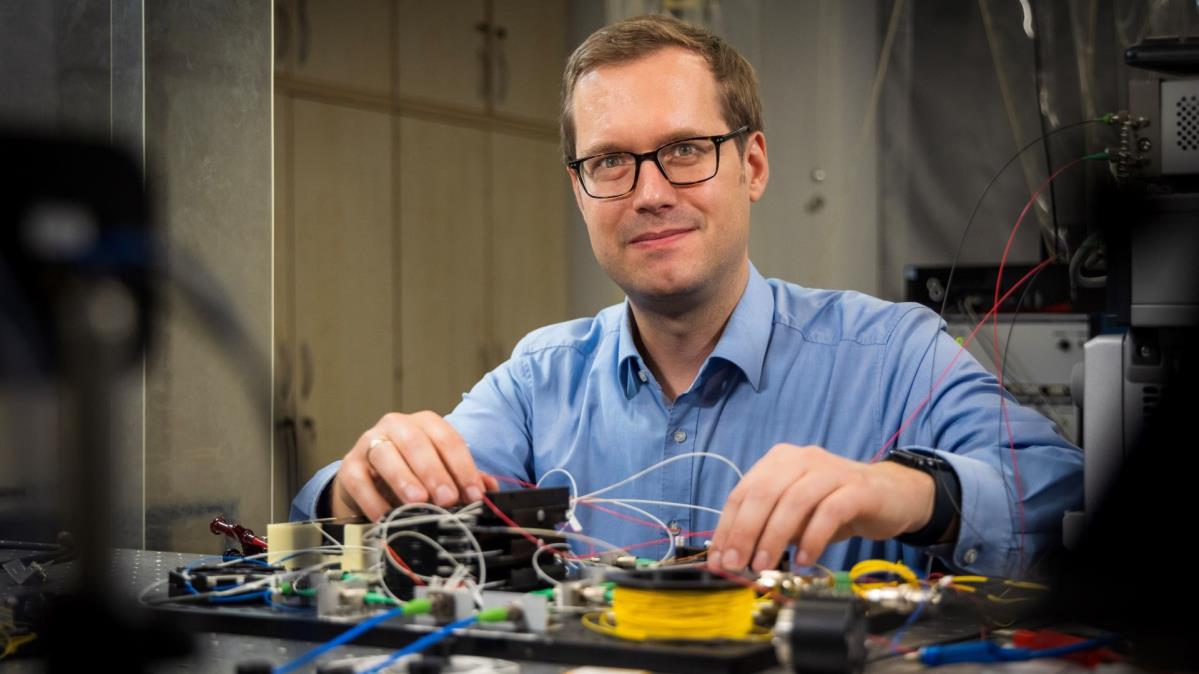 Ein Mann mit Brille und blauem Hemd sitzt an einem Labortisch mit elektronischen Komponenten und vielen Kabeln. Er blickt lächelnd in die Kamera und arbeitet konzentriert an einem Versuchsaufbau.