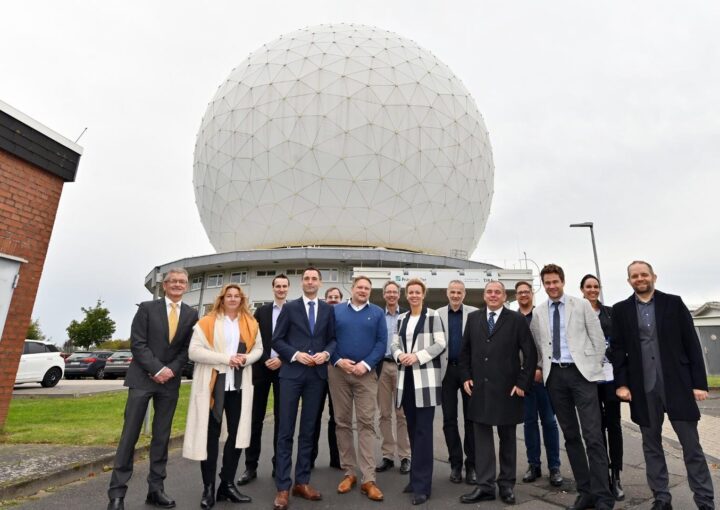 Gruppe von 14 Personen steht vor einer großen, kugelförmigen Radarkuppel mit geodätischer Struktur. Die Personen tragen überwiegend Businesskleidung und lächeln in die Kamera. Im Hintergrund ist ein Gebäude mit dem Logo des Fraunhofer-Instituts zu sehen.