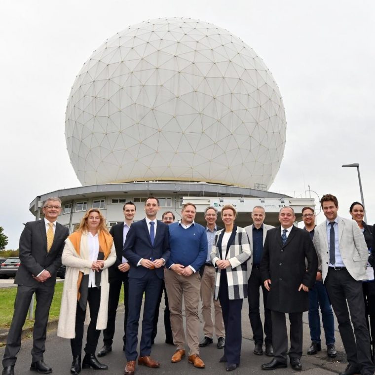 Gruppe von 14 Personen steht vor einer großen, kugelförmigen Radarkuppel mit geodätischer Struktur. Die Personen tragen überwiegend Businesskleidung und lächeln in die Kamera. Im Hintergrund ist ein Gebäude mit dem Logo des Fraunhofer-Instituts zu sehen.