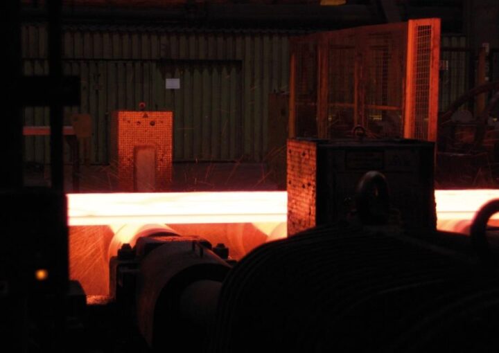 Ein glühend heißer Metallblock bewegt sich durch eine industrielle Fertigungsanlage. Funken fliegen, umgeben von Maschinen und Schutzgittern in einer dunklen Werkshalle.