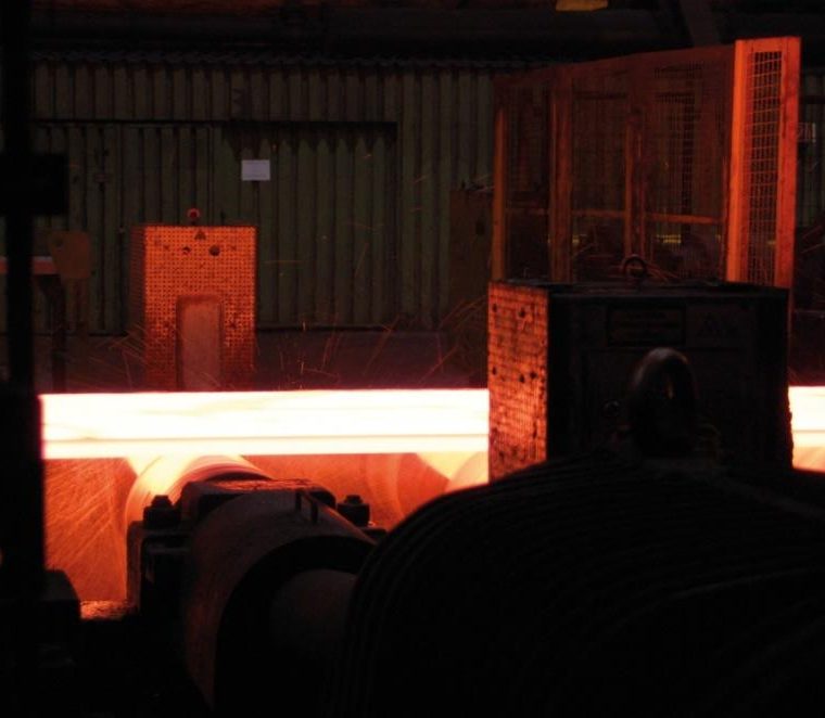Ein glühend heißer Metallblock bewegt sich durch eine industrielle Fertigungsanlage. Funken fliegen, umgeben von Maschinen und Schutzgittern in einer dunklen Werkshalle.