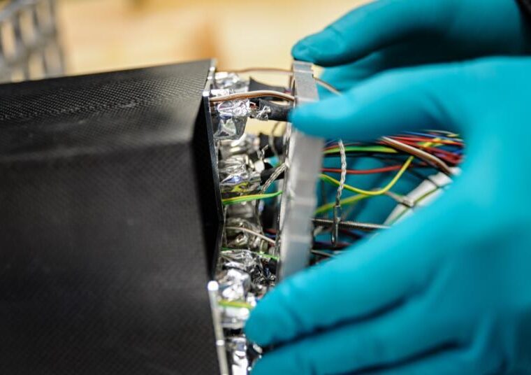 Person mit blauen Schutzhandschuhen arbeitet an einem technischen Gerät mit zahlreichen bunten Kabeln und Alufolie im Inneren. Die schwarze Gehäusestruktur wirkt technisch und präzise verarbeitet.