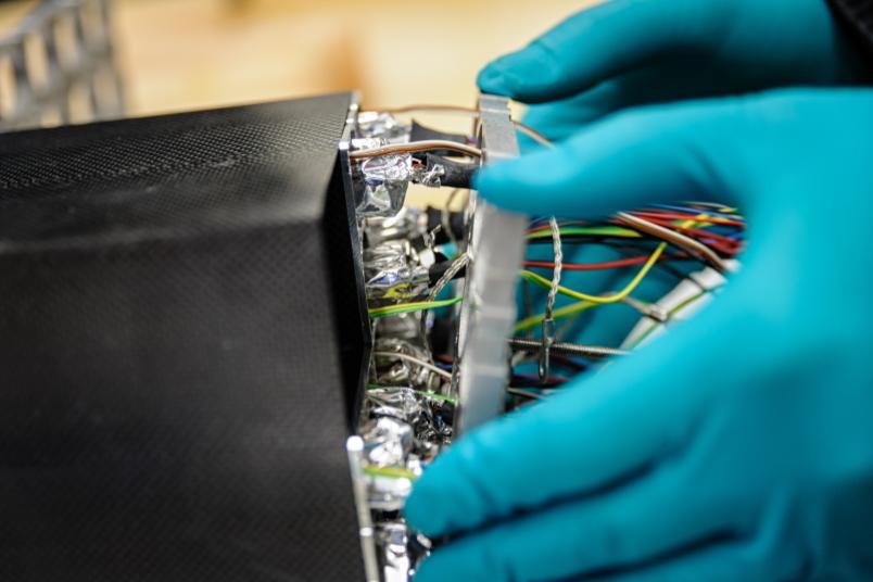 Person mit blauen Schutzhandschuhen arbeitet an einem technischen Gerät mit zahlreichen bunten Kabeln und Alufolie im Inneren. Die schwarze Gehäusestruktur wirkt technisch und präzise verarbeitet.