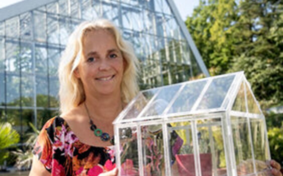 Frau steht vor einem großen Gewächshaus im Grünen und hält ein durchsichtiges Modell eines Gewächshauses in den Händen. Sie lächelt und trägt ein farbenfrohes Oberteil.