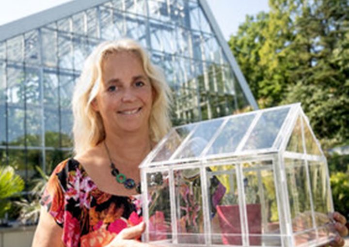 Frau steht vor einem großen Gewächshaus im Grünen und hält ein durchsichtiges Modell eines Gewächshauses in den Händen. Sie lächelt und trägt ein farbenfrohes Oberteil.