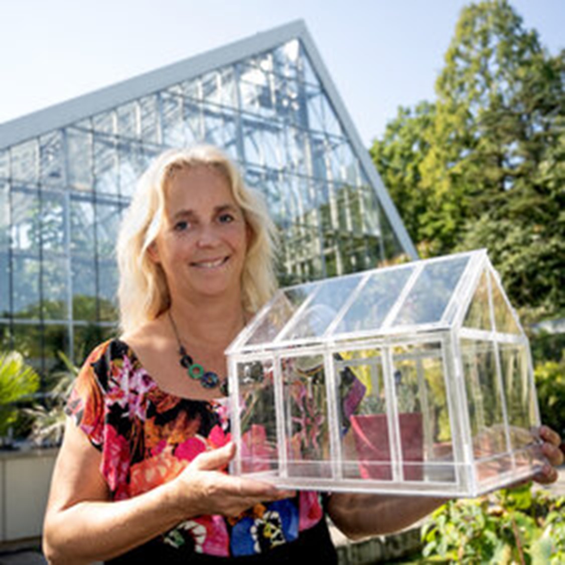 Frau steht vor einem großen Gewächshaus im Grünen und hält ein durchsichtiges Modell eines Gewächshauses in den Händen. Sie lächelt und trägt ein farbenfrohes Oberteil.