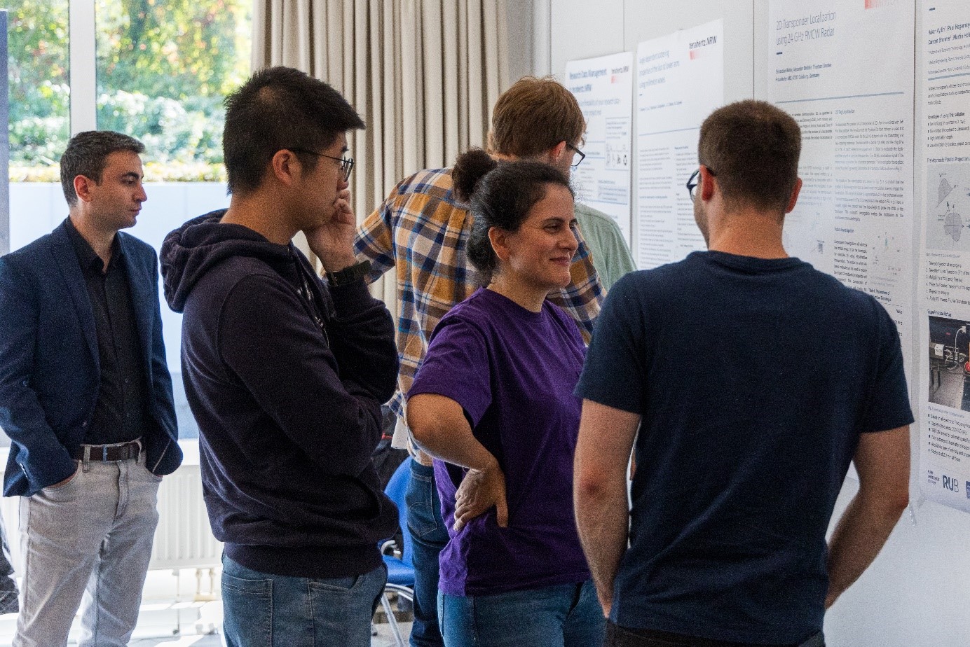 Mehrere Personen diskutieren vor wissenschaftlichen Postern. Im Vordergrund unterhält sich eine Frau im lila Shirt angeregt mit anderen Teilnehmenden.