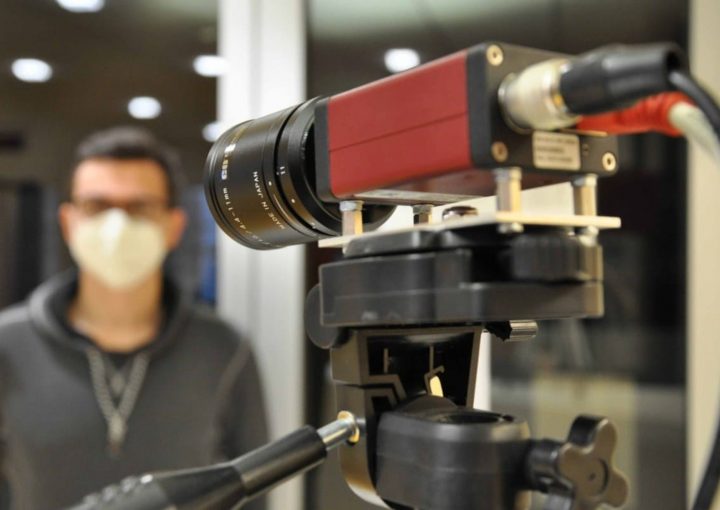 Im Vordergrund steht eine wissenschaftliche Messkamera mit Objektiv auf einem Stativ. Im Hintergrund ist eine Person mit FFP2-Maske unscharf zu erkennen, offenbar Teil eines Experiments oder einer Messung.