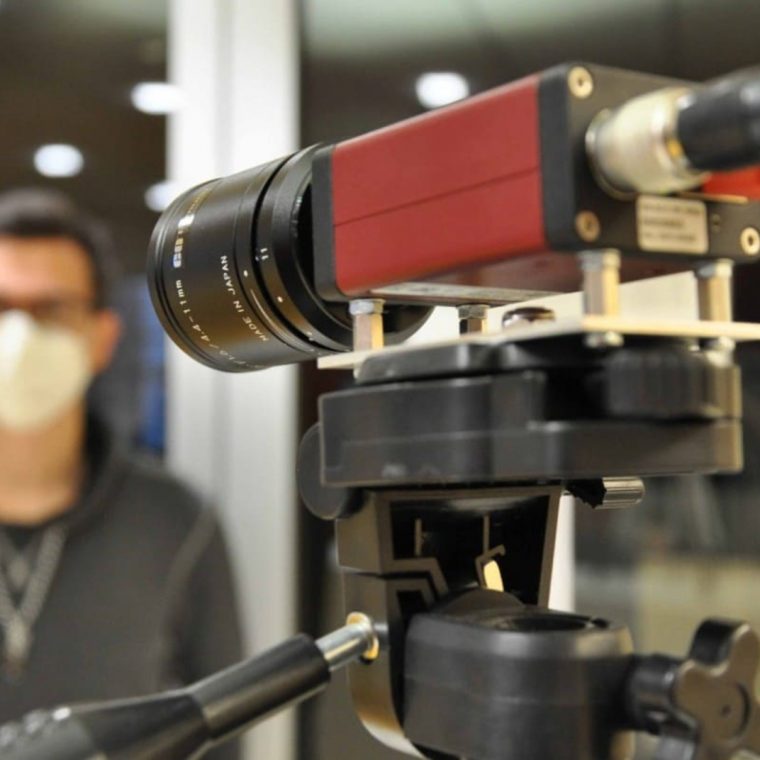 Im Vordergrund steht eine wissenschaftliche Messkamera mit Objektiv auf einem Stativ. Im Hintergrund ist eine Person mit FFP2-Maske unscharf zu erkennen, offenbar Teil eines Experiments oder einer Messung.