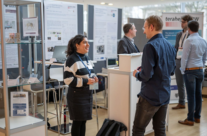 Mehrere Personen im Gespräch an Messeständen mit Postern und Vitrinen auf einer wissenschaftlichen Konferenz. Im Vordergrund unterhalten sich eine Frau und ein Mann freundlich.