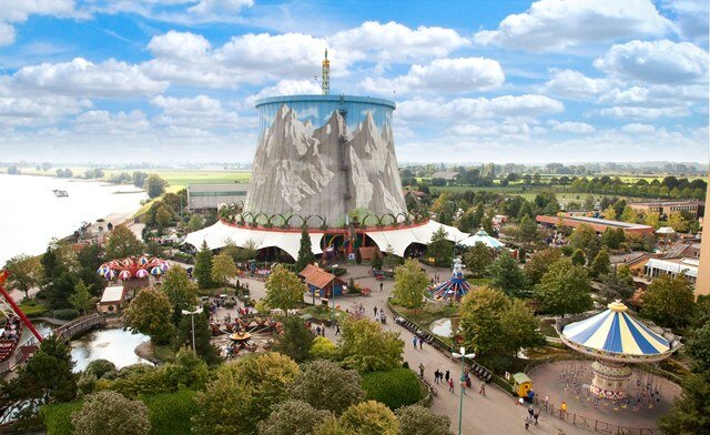 Luftaufnahme eines Freizeitparks mit zentralem, bergbemaltem Turm. Umgeben von Karussells, Grünflächen, Wegen und Besuchergruppen. Im Hintergrund ein Fluss und weite Landschaft.